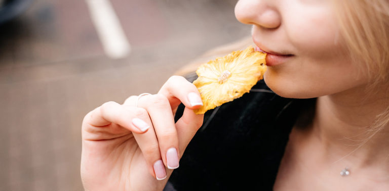 Una donna bionda con una giacca di pelle nera e unghie laccate di rosa tiene un pezzo di ananas essiccato vicino alle labbra per mangiarlo.
