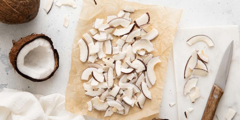 Composizione dall'alto che mostra delle scaglie di cocco essiccato e tostato disposte su carta da forno al centro, con mezzi cocchi freschi e un coltello da cucina con manico in legno sui lati, su uno sfondo bianco e grigio marmorizzato