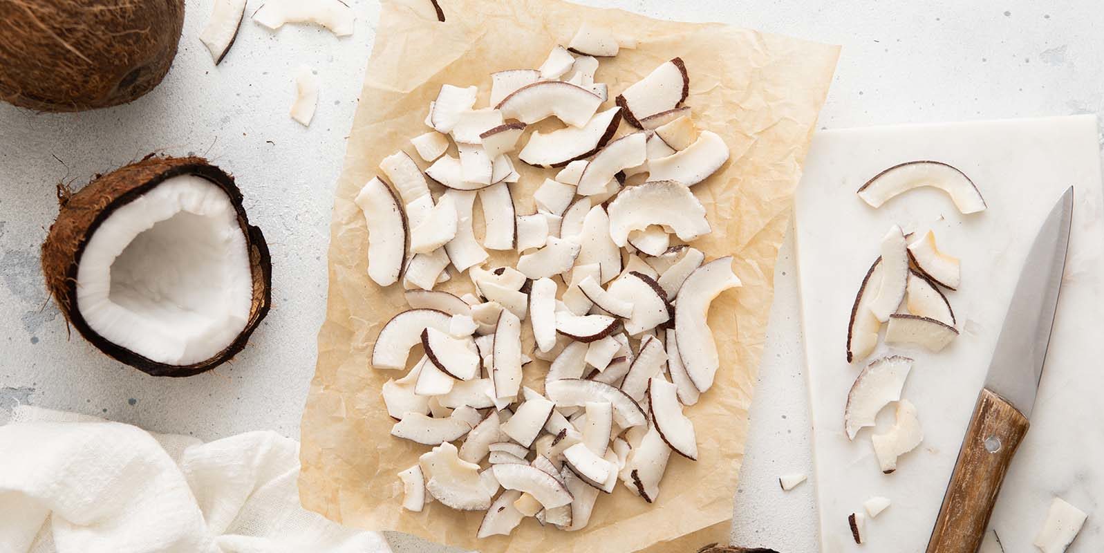 Composizione dall'alto che mostra delle scaglie di cocco essiccato e tostato disposte su carta da forno al centro, con mezzi cocchi freschi e un coltello da cucina con manico in legno sui lati, su uno sfondo bianco e grigio marmorizzato