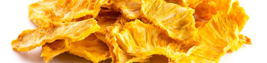 Un mucchio di sottili e raggrinziti pezzi di ananas essiccata, di un brillante colore giallo-dorato, è sparso su uno sfondo bianco. L'ananas essiccato conserva le sue numerose proprietà benefiche.