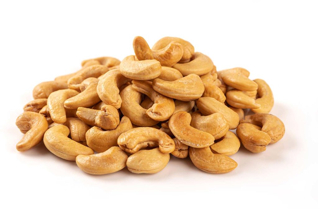 Pila isolata di anacardi interi tostati, dalla forma a mezzaluna, di colore dorato chiaro, su uno sfondo bianco neutro.