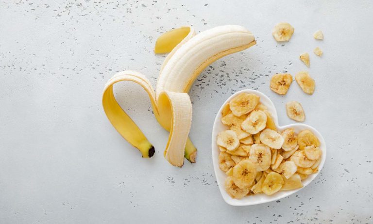Una banana sbucciata parzialmente accanto a una ciotola bianca a forma di cuore piena di fette di banana essiccata.