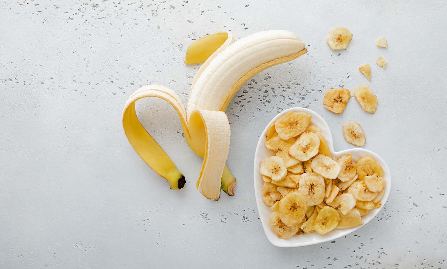 Una banana sbucciata parzialmente accanto a una ciotola bianca a forma di cuore piena di fette di banana essiccata.