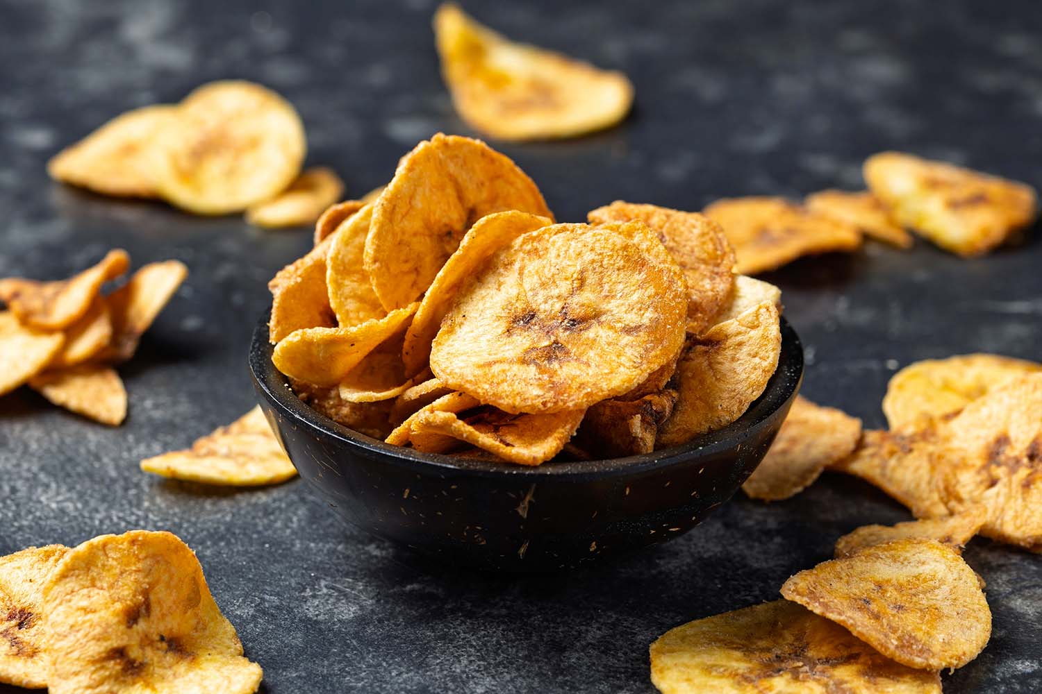 Una ciotola di cocco scura piena di chips di banana essiccata e croccante, su uno sfondo ruvido e scuro.