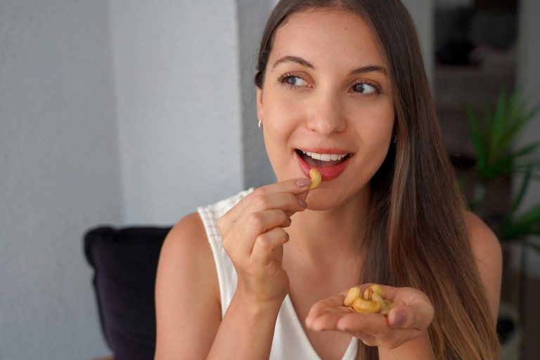 Ritratto ravvicinato di una donna con lunghi capelli castani, vestita con una canotta bianca, che porta un anacardo alla bocca con un'espressione felice e soddisfatta. Nella sua mano sinistra tiene una piccola manciata di anacardi.