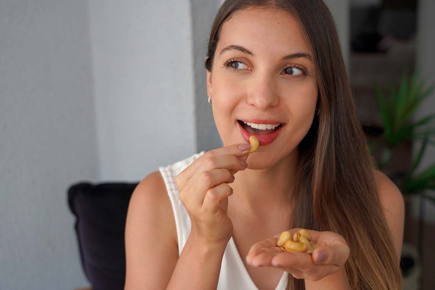 Ritratto ravvicinato di una donna con lunghi capelli castani, vestita con una canotta bianca, che porta un anacardo alla bocca con un'espressione felice e soddisfatta. Nella sua mano sinistra tiene una piccola manciata di anacardi.