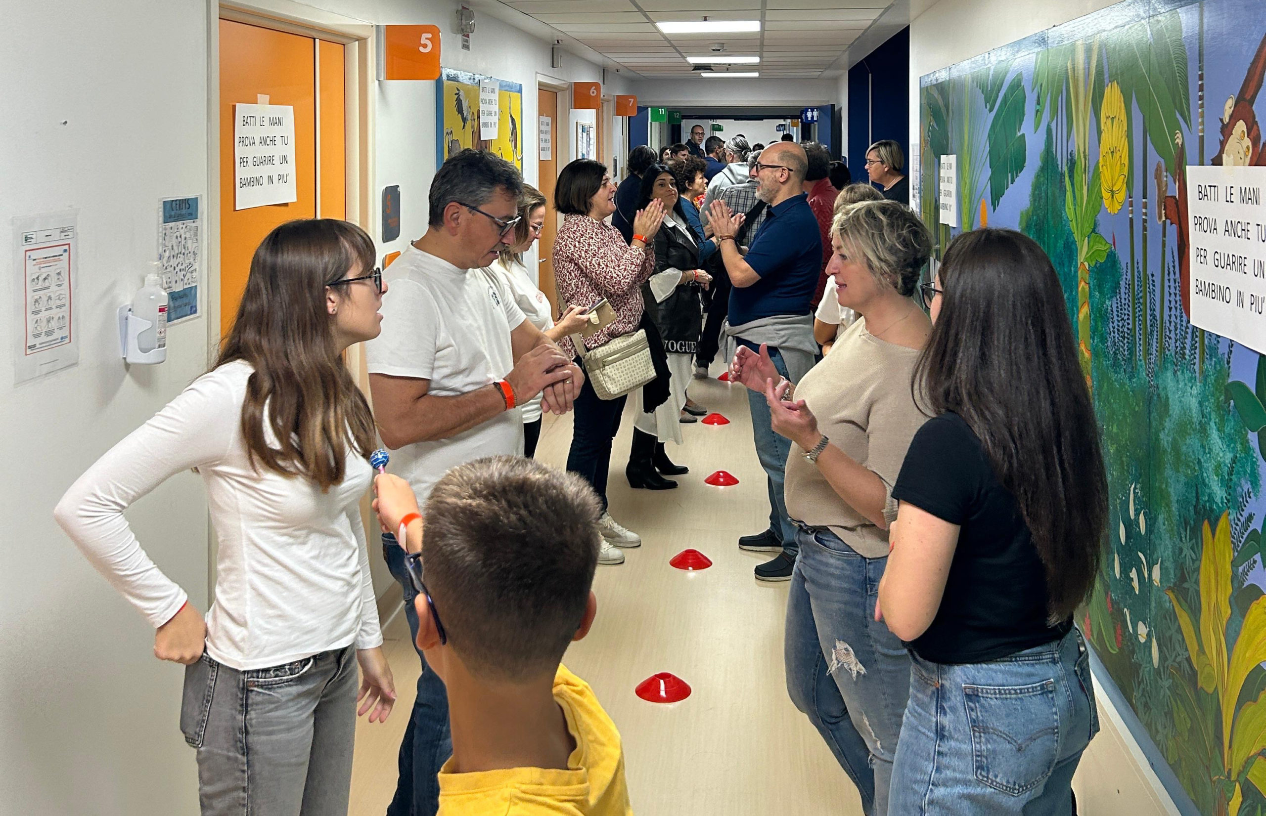 Partecipanti e volontari si incontrano in un corridoio colorato del Centro Maria Letizia Verga. Si vedono persone in conversazione in fila, con segnaposto rossi sul pavimento e il cartello del motto 'Batti le mani prova anche tu...' in evidenza sulla parete decorata con murales naturalistici, in attesa del tentativo di Guinness World Record