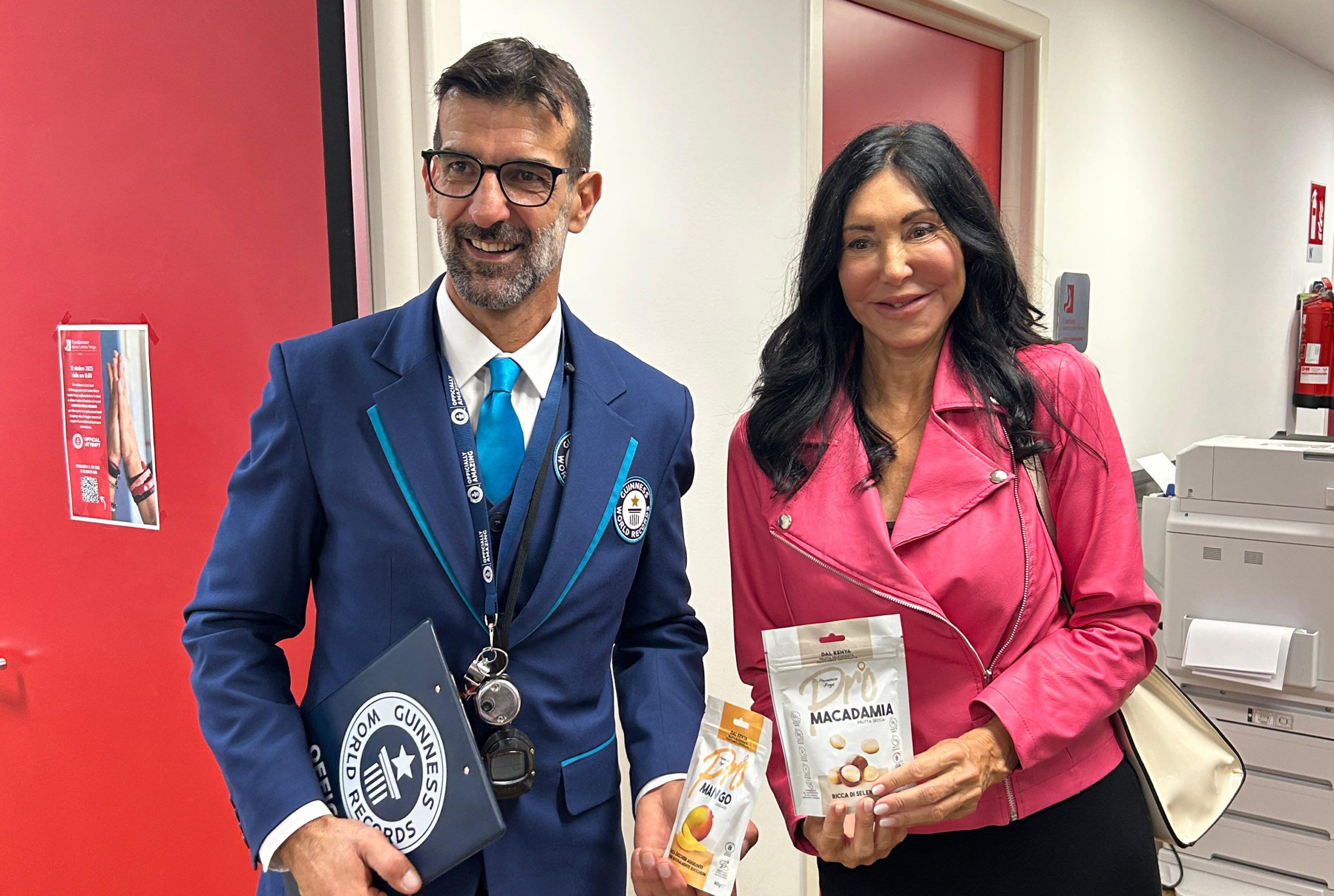 Il giudice ufficiale del Guinness World Records in abito blu con distintivo e Giulia Galli, CEO di Dracma Educational, che mostra due confezioni di frutta secca o snack Pro. Scena scattata all'interno del Centro Maria Letizia Verga durante l'evento record.