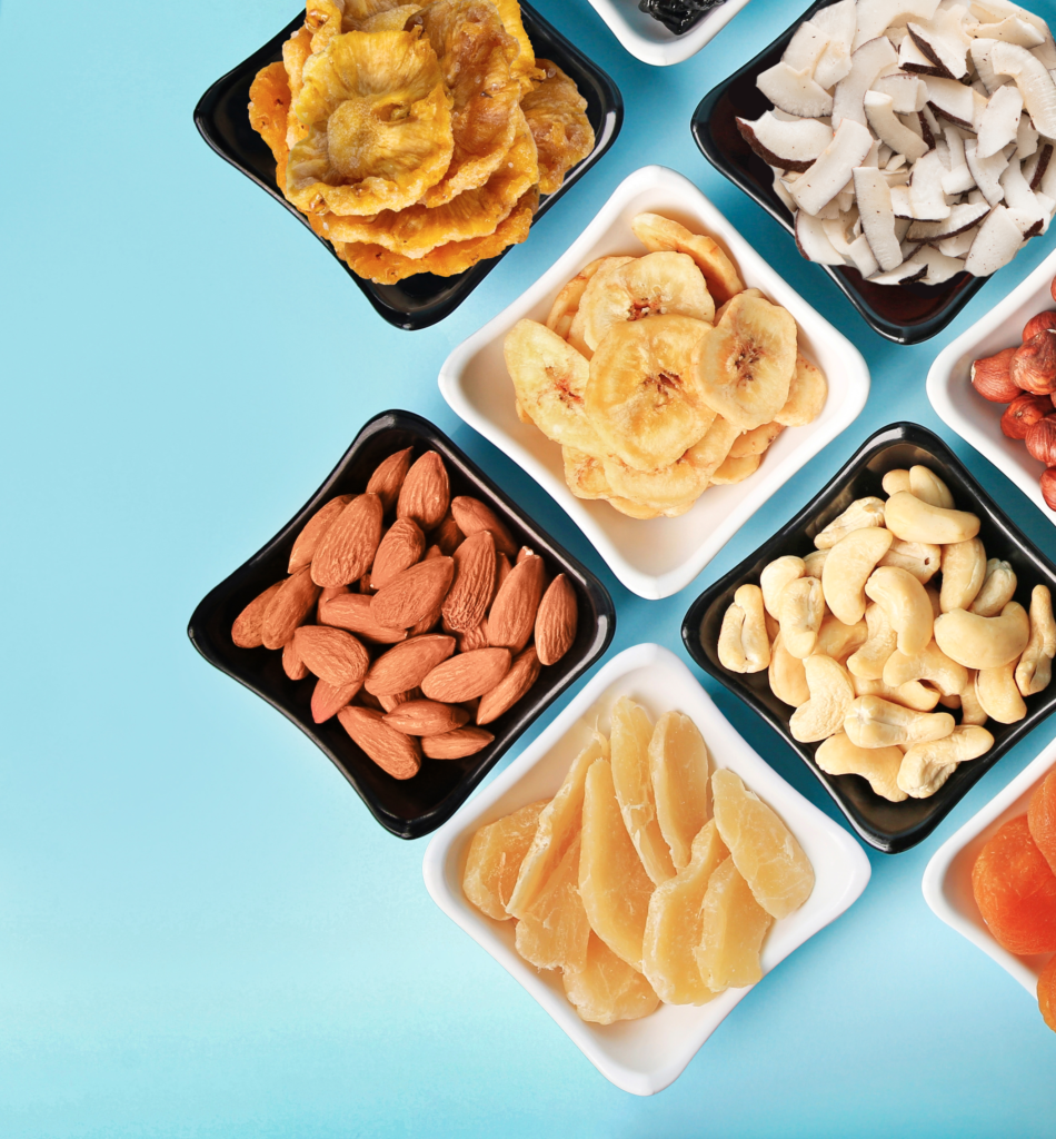 Vari tipi di frutta secca e frutta essiccata disposti in piccole ciotole quadrate bianche e nere su uno sfondo blu chiaro. Si distinguono mandorle, anacardi, cocco a scaglie, ananas essiccato e banana essiccata. Mix di snack salutari, vegani, senza glutine, ingredienti per colazione, cibo energetico.