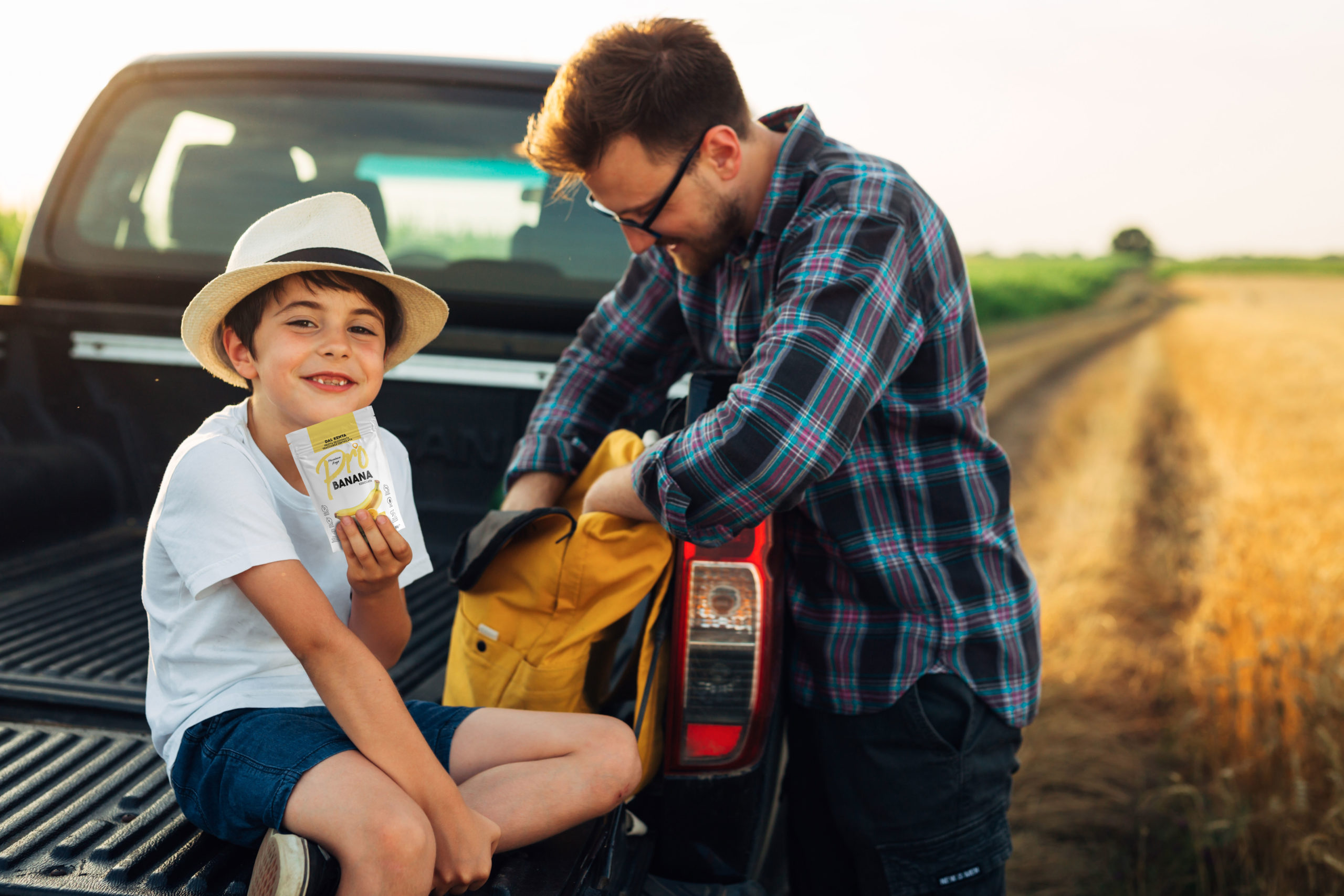 Un padre e suo figlio seduti sul portellone posteriore di un pick-up in un campo di grano al tramonto. Il padre con occhiali e camicia a quadri sistema uno zaino giallo, mentre il figlio con cappello di paglia e maglietta bianca sorride alla telecamera, mostrando una confezione di snack di frutta secca monoporzione Pro alla banana. Scena rurale, famiglia, snack, viaggio, agricoltura, natura.